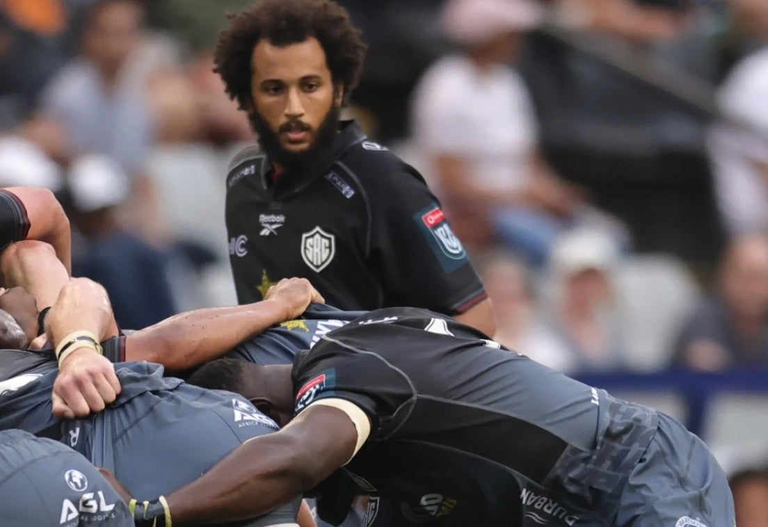 A Hollywoodbets Sharks player in a black jersey watches over a scrum as the forwards bind, with the crowd blurred in the stands behind.