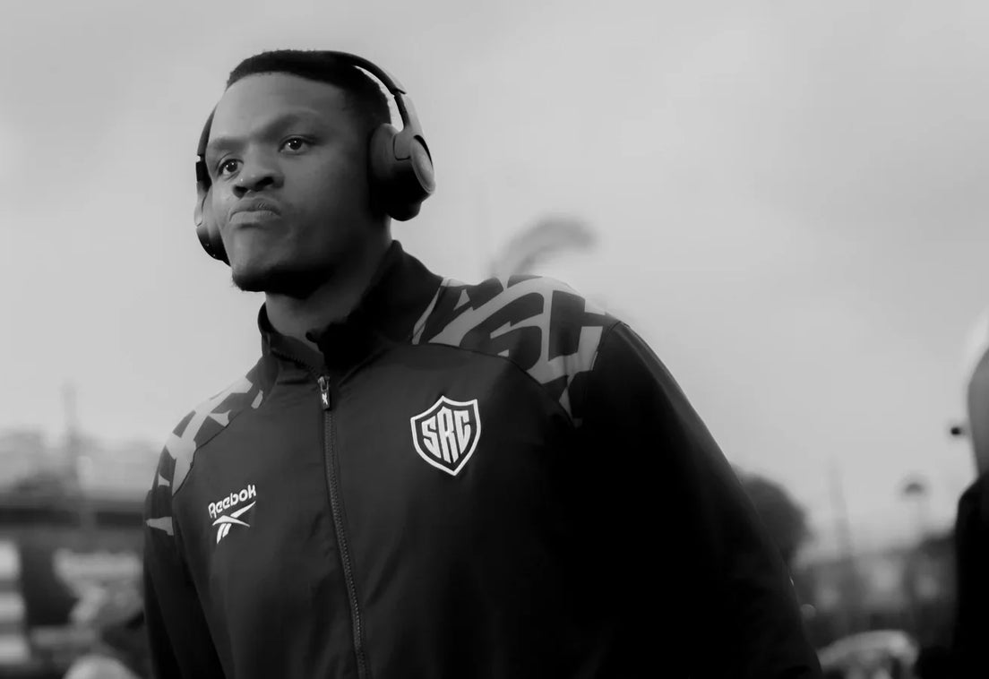 A Hollywoodbets Sharks player wearing headphones and a black Sharks tracksuit pulls a travel bag as he arrives at the stadium on an overcast day.