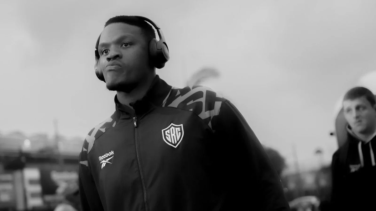 A Hollywoodbets Sharks player wearing headphones and a black Sharks tracksuit pulls a travel bag as he arrives at the stadium on an overcast day.