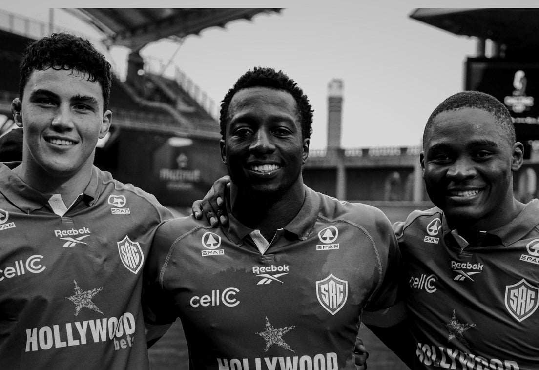 Three smiling young Sharks players stand arm in arm in their black jerseys inside an empty stadium, in a black and white photo.