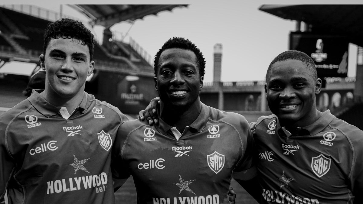 Three smiling young Sharks players stand arm in arm in their black jerseys inside an empty stadium, in a black and white photo.