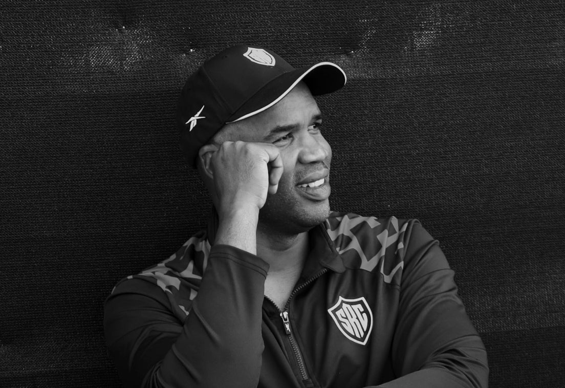 Black and white photo of Sharks head coach JP Pietersen in a Sharks cap and training jacket, smiling as he looks off to one side.