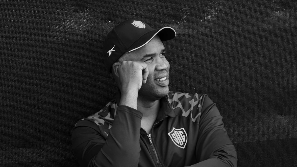 Black and white photo of Sharks head coach JP Pietersen in a Sharks cap and training jacket, smiling as he looks off to one side.
