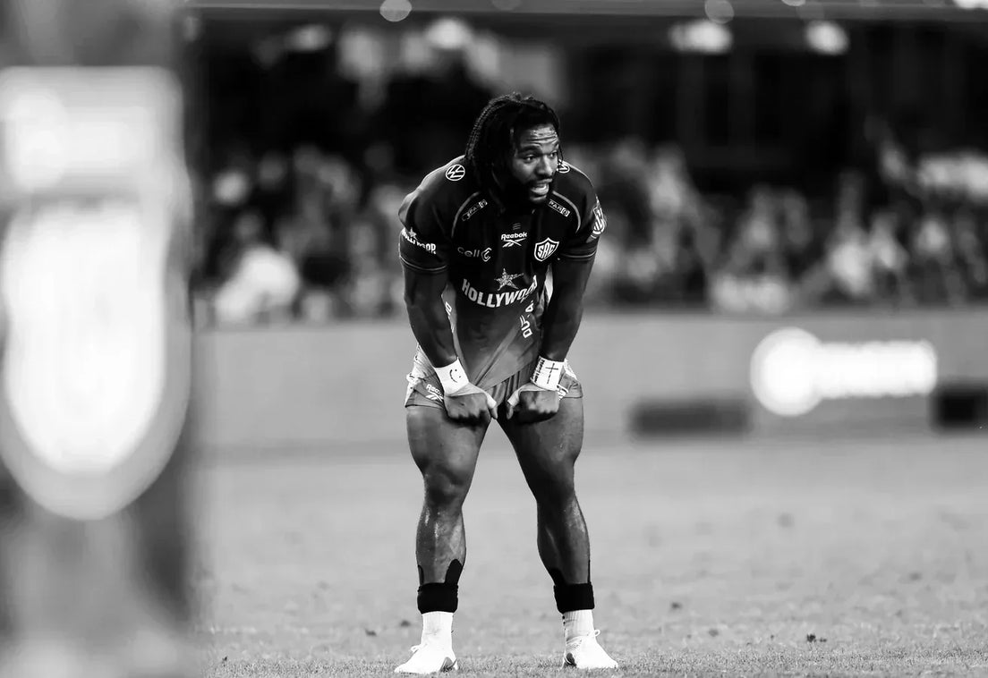 Black-and-white photo of a Hollywoodbets Sharks player with long dreadlocks, hands on his knees catching his breath on the pitch, with a packed crowd blurred behind him.