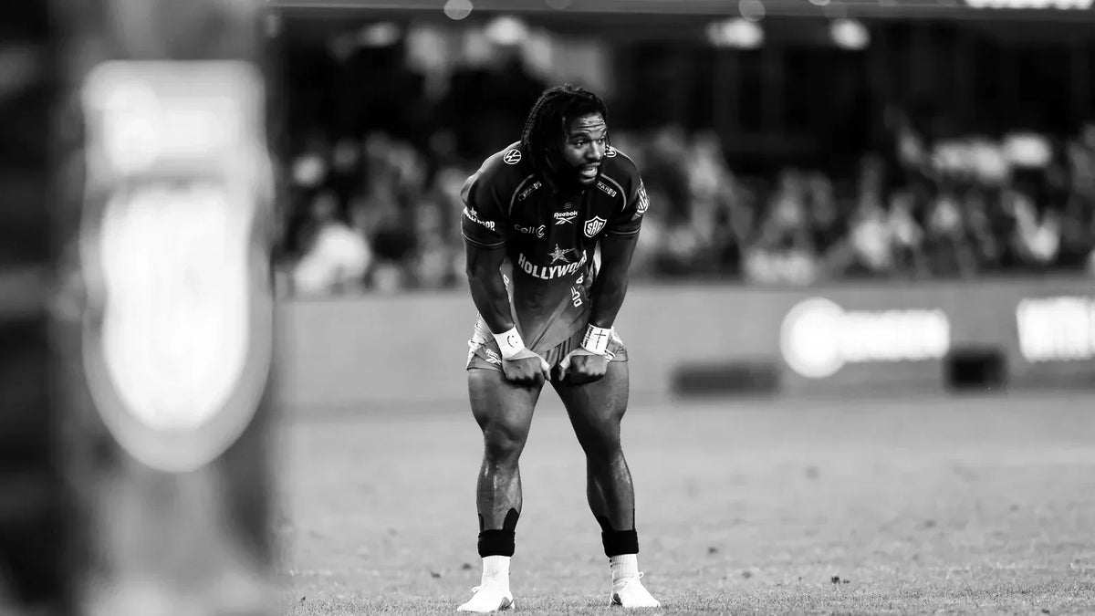 Black-and-white photo of a Hollywoodbets Sharks player with long dreadlocks, hands on his knees catching his breath on the pitch, with a packed crowd blurred behind him.