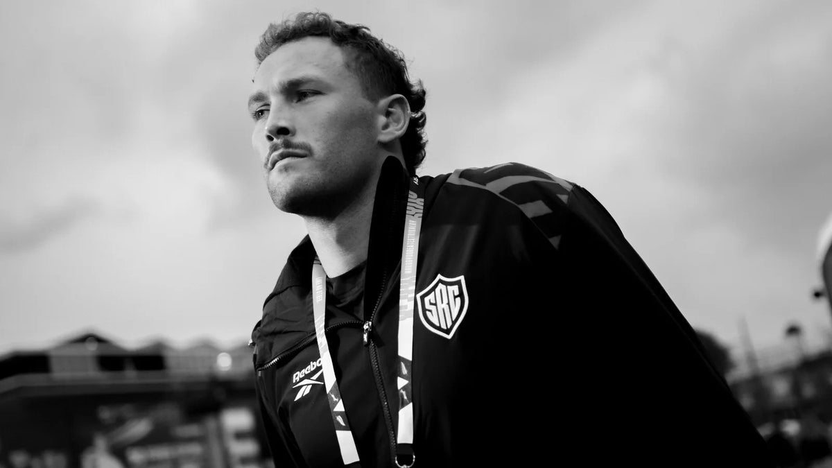 Black-and-white photo of Nick Hatton in a black team jacket and match accreditation lanyard, walking under a cloudy sky.