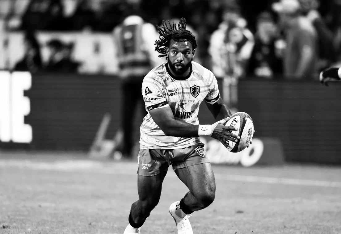 A Sharks player in a training top runs onto the ball with it held in both hands during a floodlit pre-match warm-up.