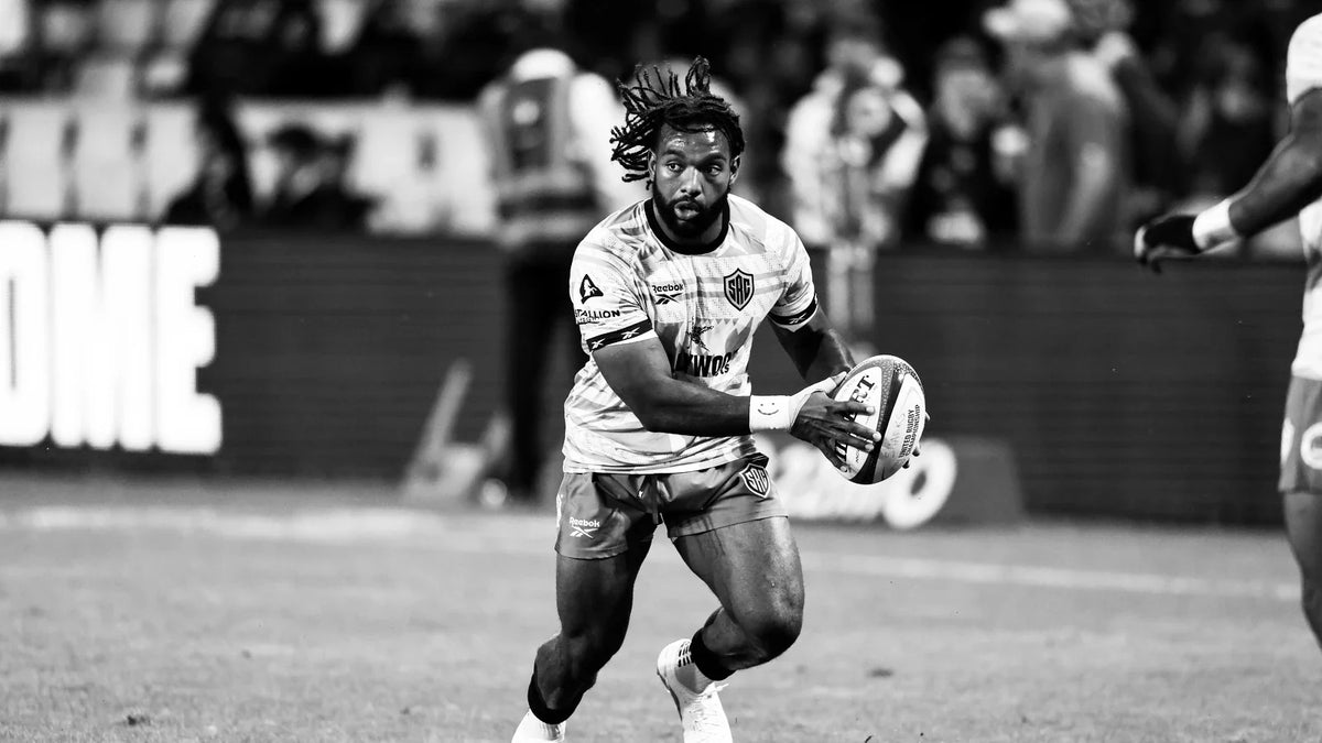 A Sharks player in a training top runs onto the ball with it held in both hands during a floodlit pre-match warm-up.