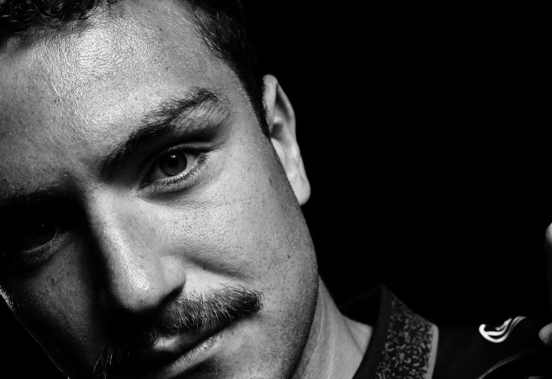 Black and white close-up portrait of Janco Purchase with a curly fringe and moustache, head tilted, tugging at the collar of his Hollywoodbets Sharks jersey against a black background.
