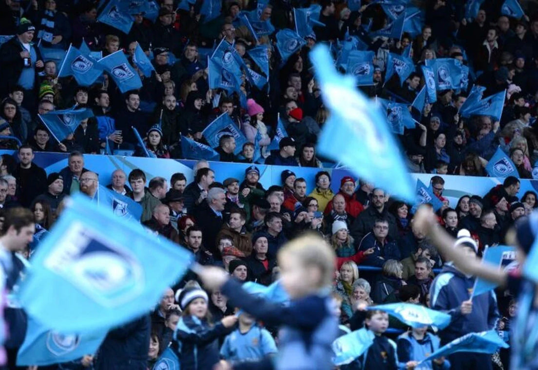 Cardiff Rugby supporters, young and old, wave light blue club flags in a packed home stand, with children in the foreground.