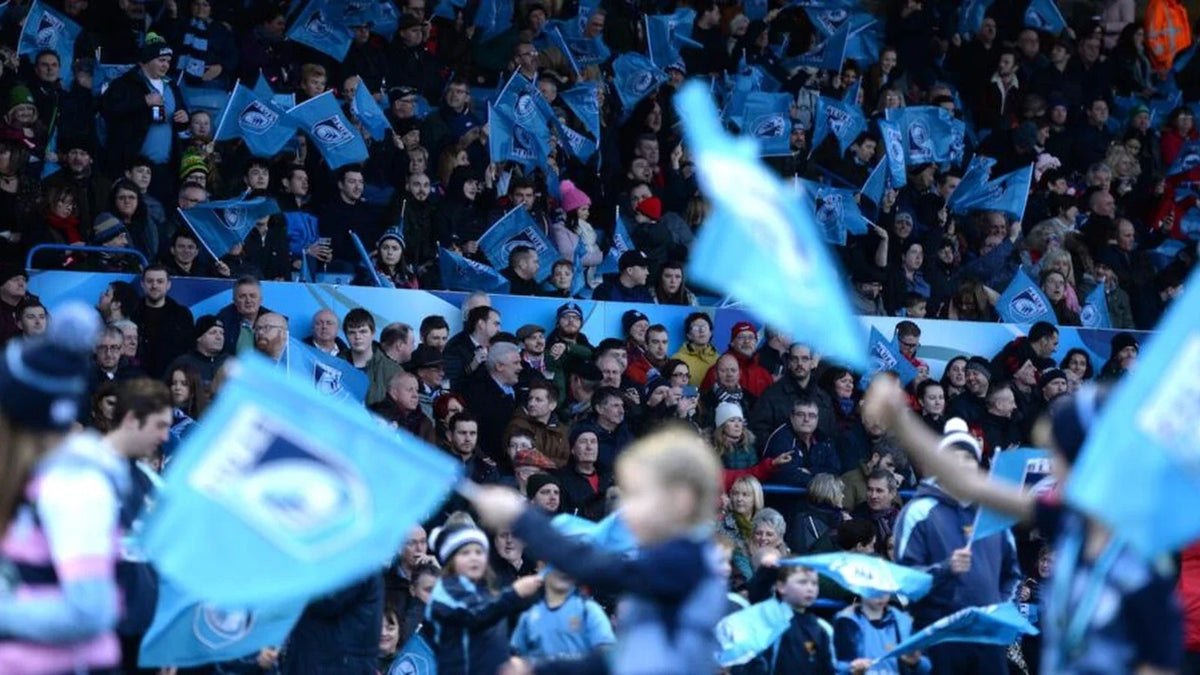 Cardiff Rugby supporters, young and old, wave light blue club flags in a packed home stand, with children in the foreground.