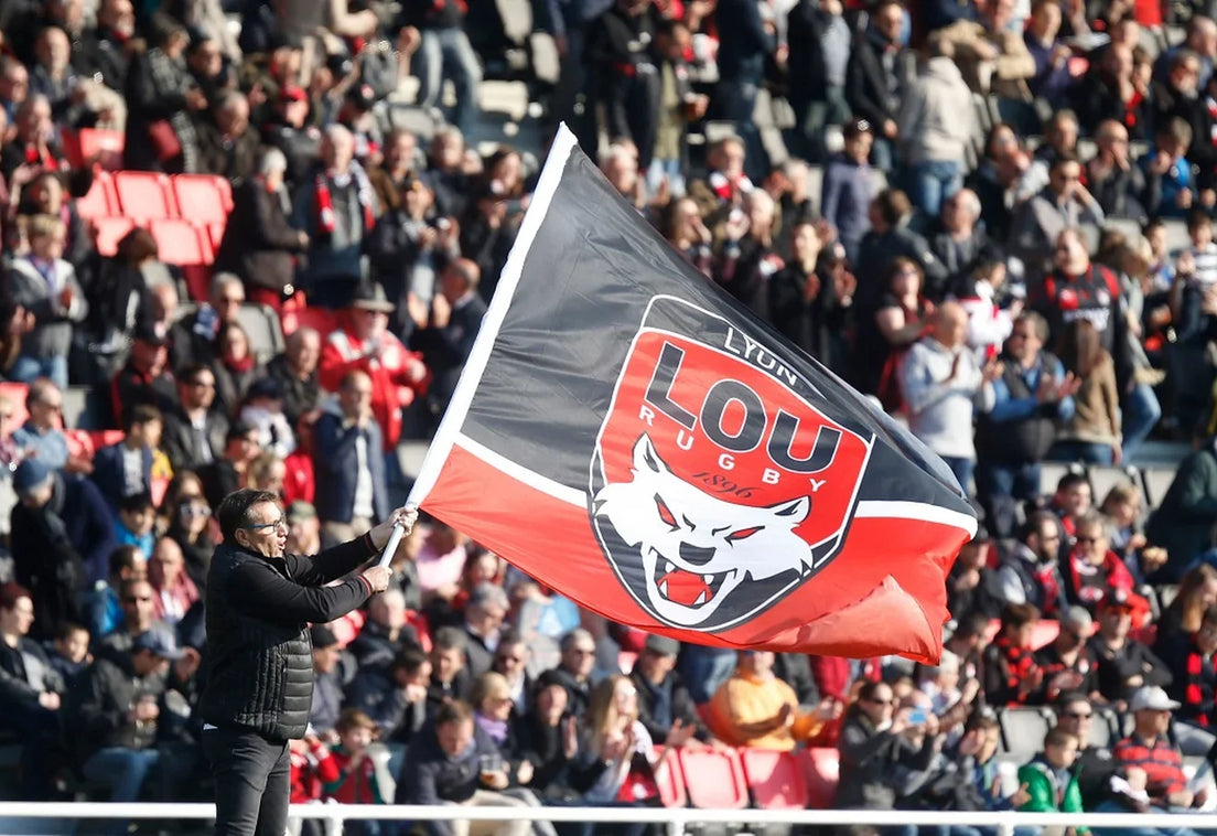 A Lyon supporter waves a large red and black LOU Rugby flag in front of a packed stand of fans in red and black.