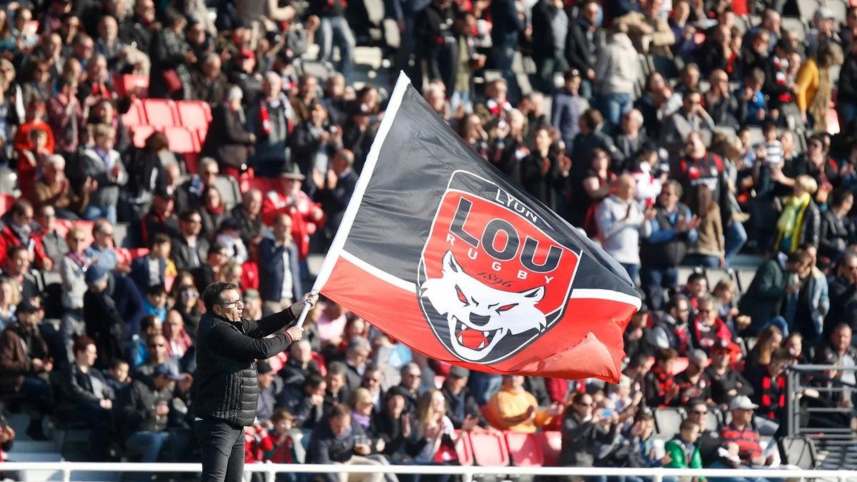 A Lyon supporter waves a large red and black LOU Rugby flag in front of a packed stand of fans in red and black.