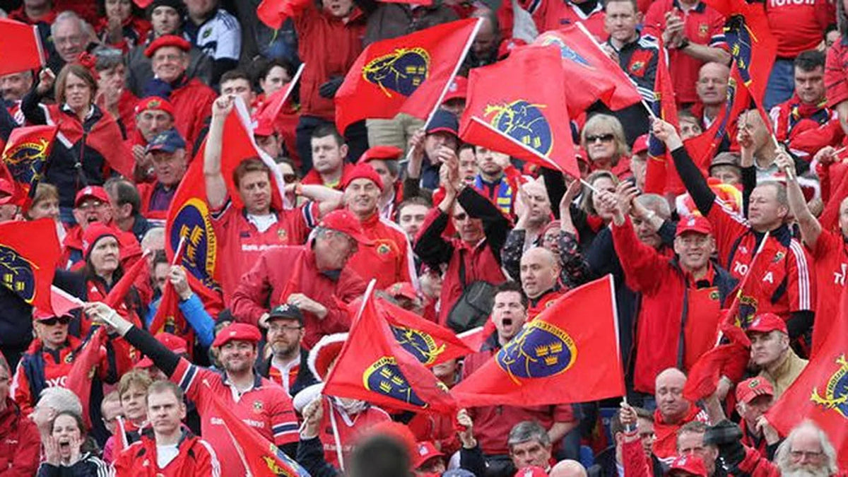 Munster supporters in red caps and jerseys wave red flags bearing the province's crest across a packed, cheering stand.