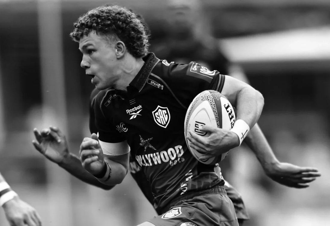 A Hollywoodbets Sharks player sprints with the ball tucked under his right arm as a team-mate tracks in support, in black and white.
