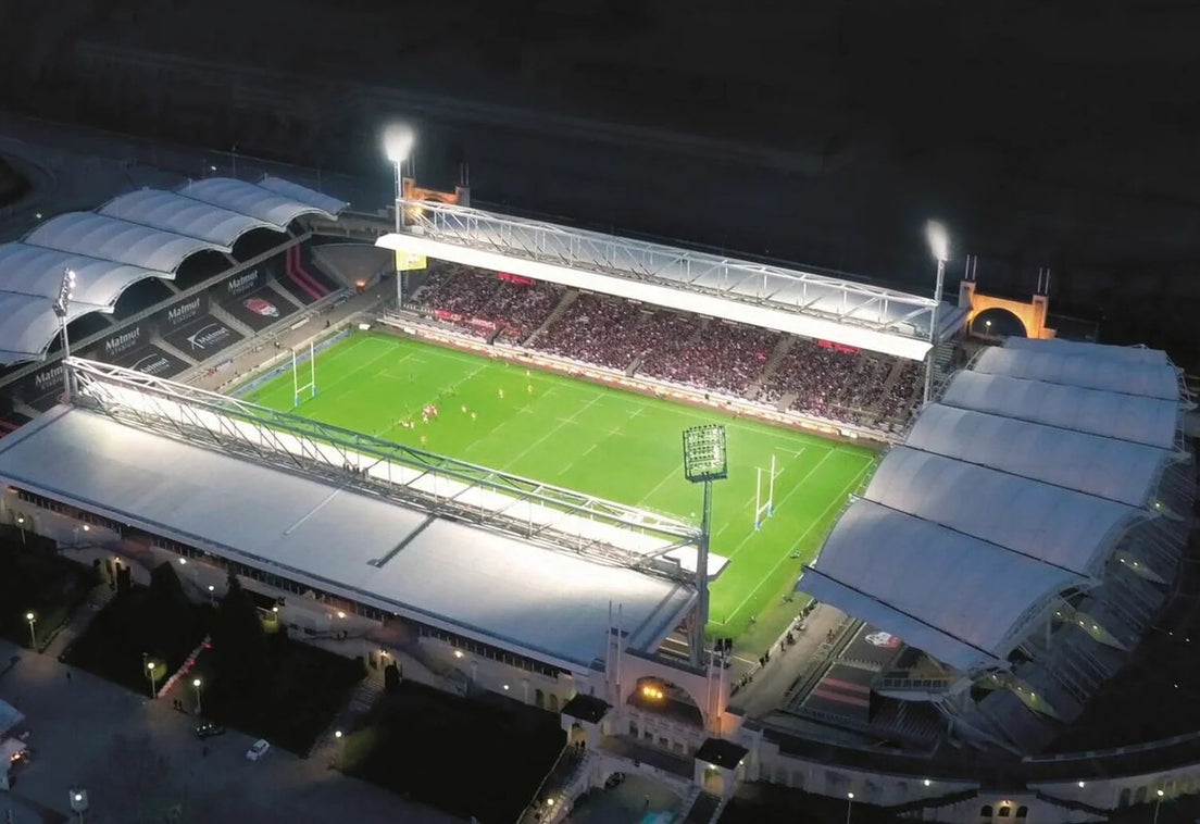 Aerial night view of Lyon's floodlit Matmut Stadium de Gerland, with a rugby match under way on the green pitch in front of a packed main stand.