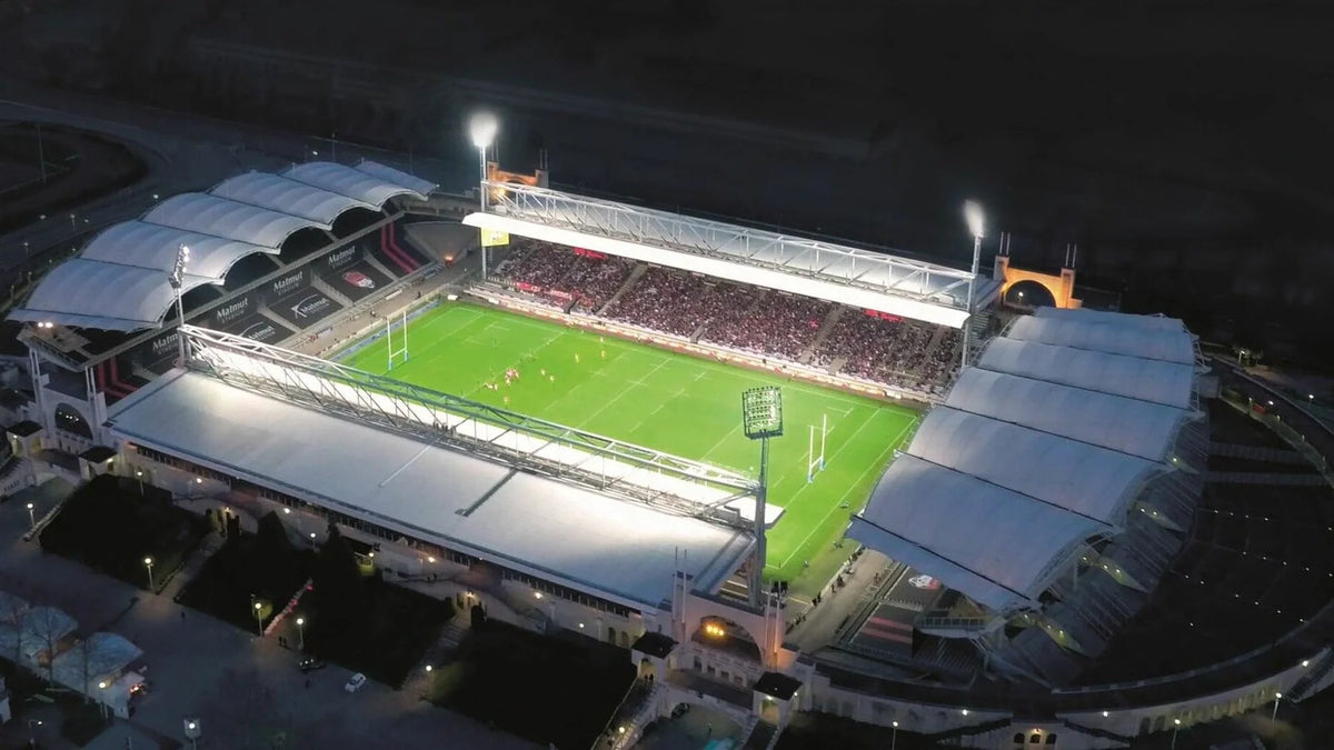 Aerial night view of Lyon's floodlit Matmut Stadium de Gerland, with a rugby match under way on the green pitch in front of a packed main stand.
