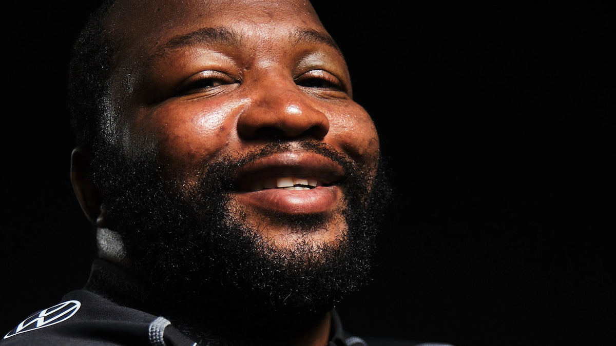 Close-up of a smiling, bearded Hollywoodbets Sharks prop in the black home jersey, lit dramatically against a black background.