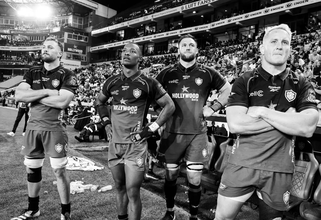 Five Sharks players stand on the touchline at Hollywoodbets Kings Park with arms folded and hands on hips, watching the game under the floodlights.