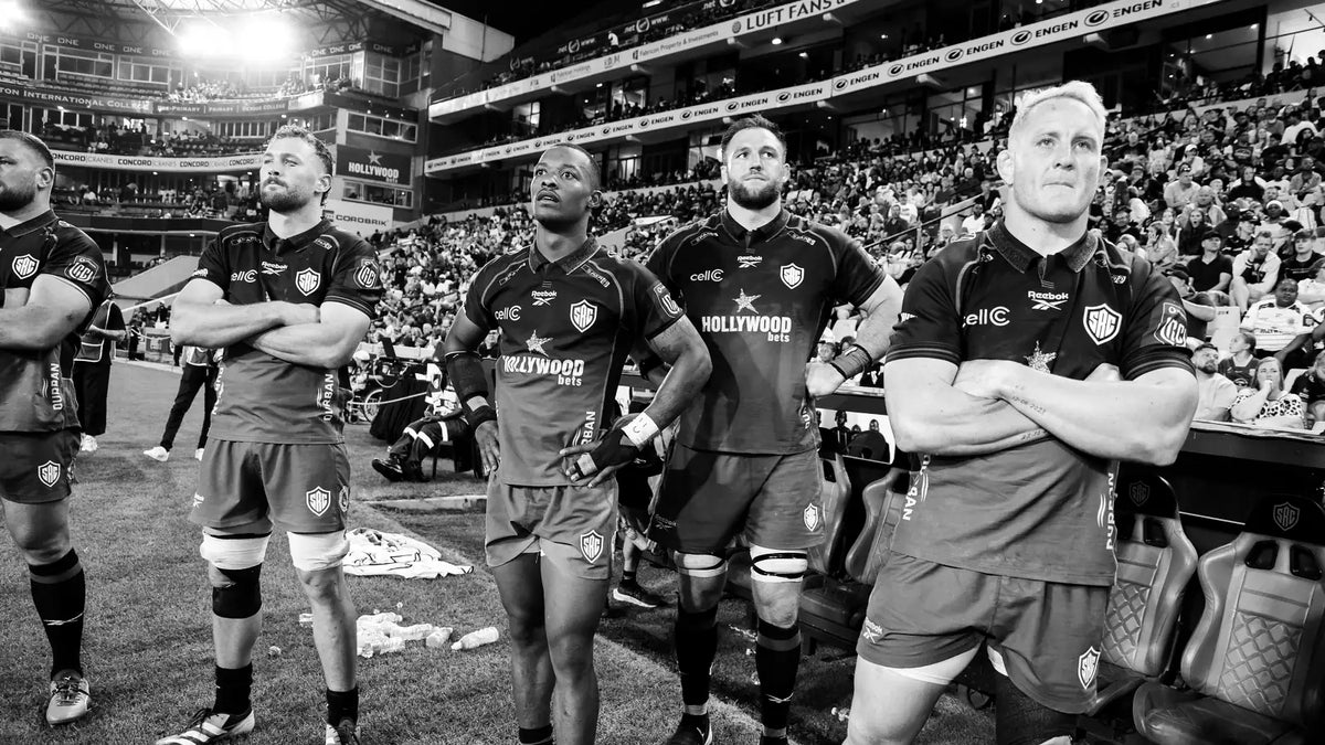 Five Sharks players stand on the touchline at Hollywoodbets Kings Park with arms folded and hands on hips, watching the game under the floodlights.