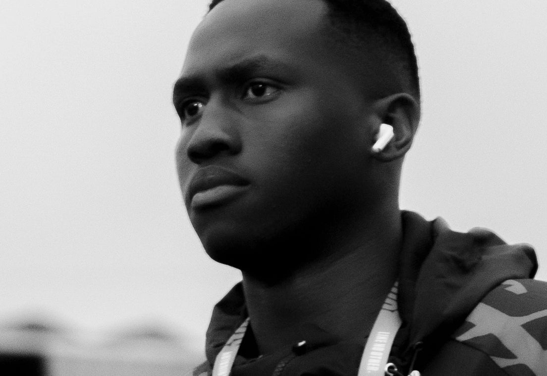 Black and white close-up of a Hollywoodbets Sharks player in a club rain jacket and accreditation lanyard, wearing an earbud and looking off to one side against an overcast sky.