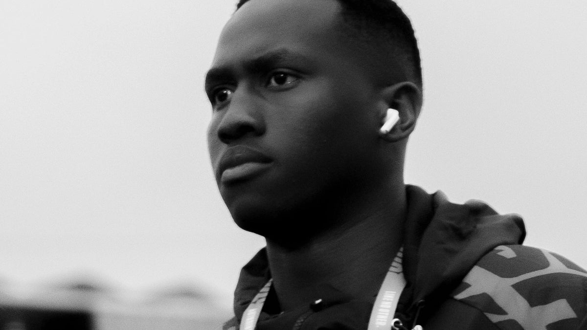 Black and white close-up of a Hollywoodbets Sharks player in a club rain jacket and accreditation lanyard, wearing an earbud and looking off to one side against an overcast sky.