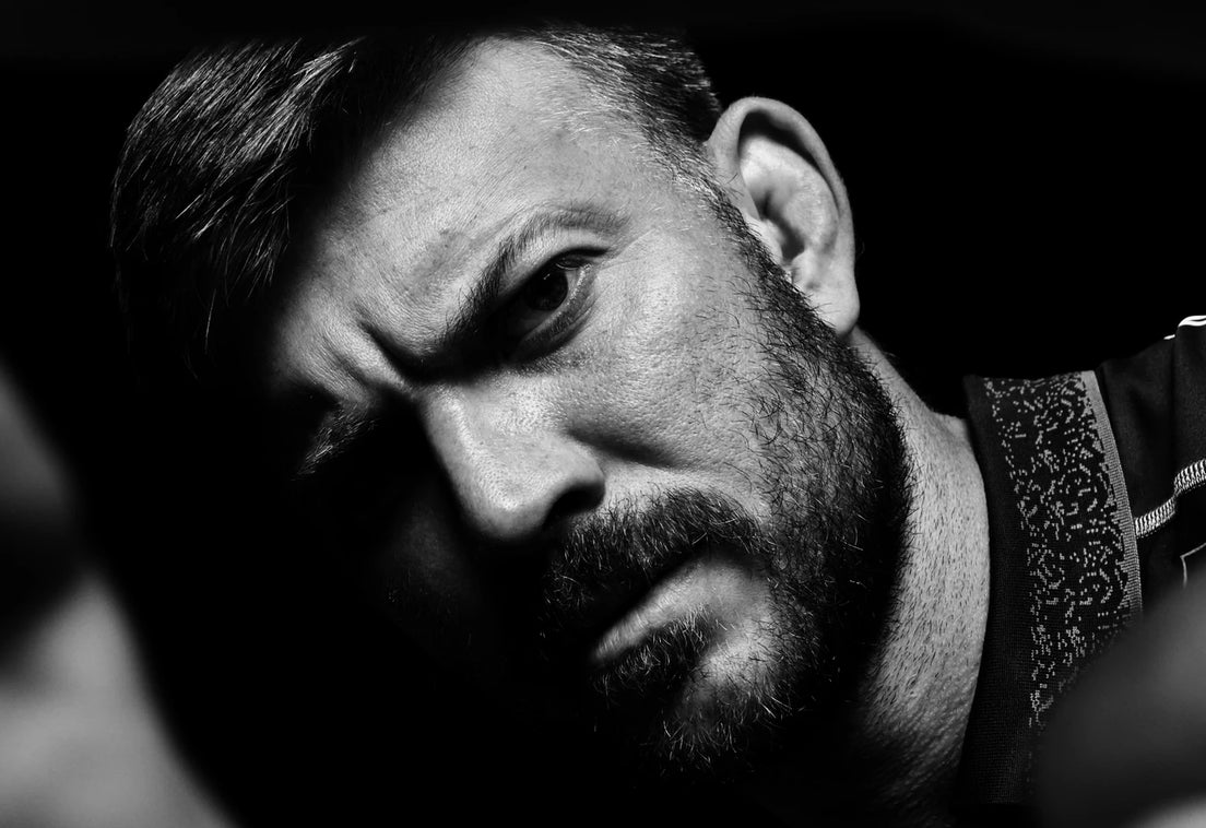 A bearded man in a black Sharks training shirt frowns into the camera through his outstretched hands in a black and white studio portrait.