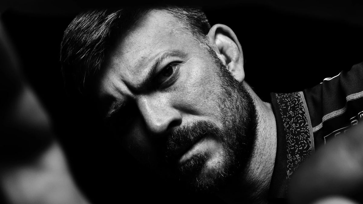 A bearded man in a black Sharks training shirt frowns into the camera through his outstretched hands in a black and white studio portrait.