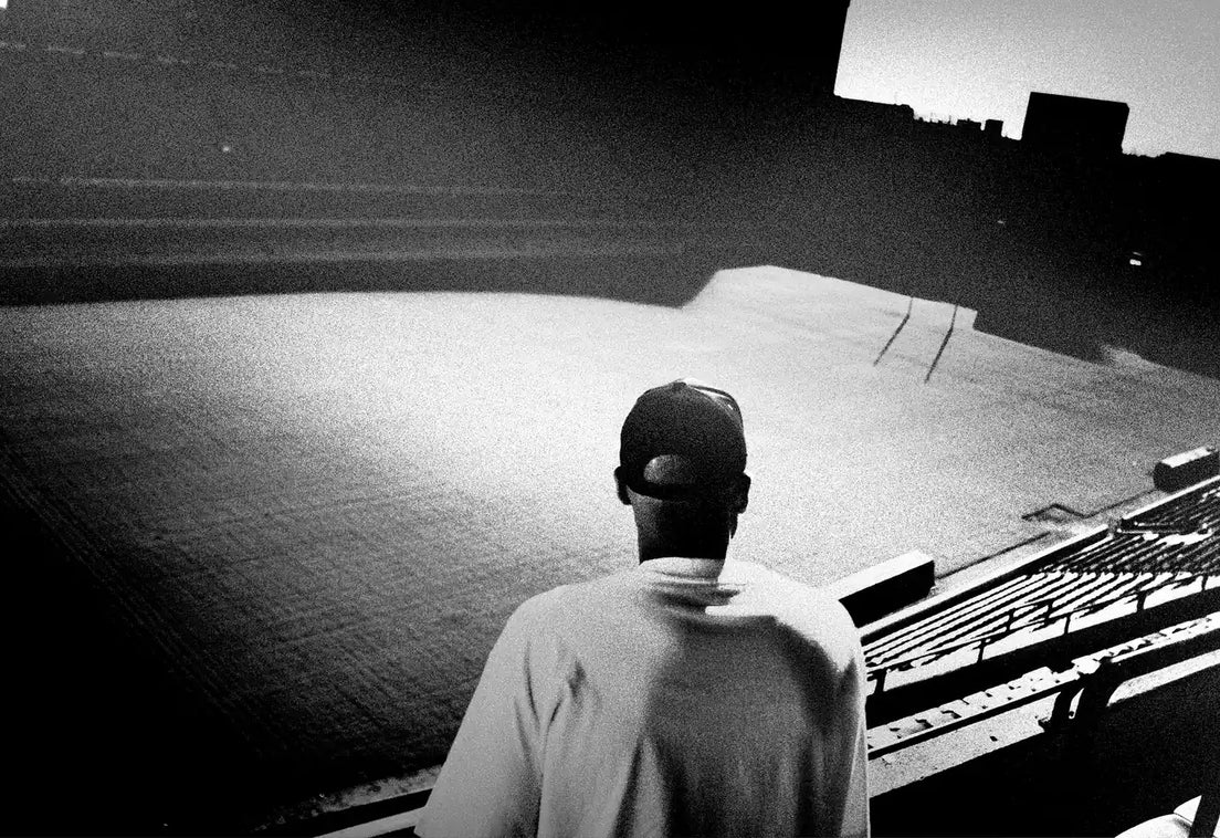 A lone figure in a cap and pale top stands in the empty stands at dusk, looking out over a deserted rugby pitch and goalposts