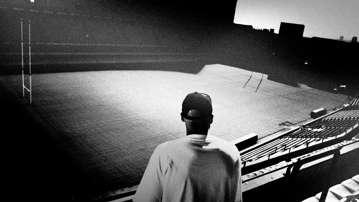 A lone figure in a cap and pale top stands in the empty stands at dusk, looking out over a deserted rugby pitch and goalposts
