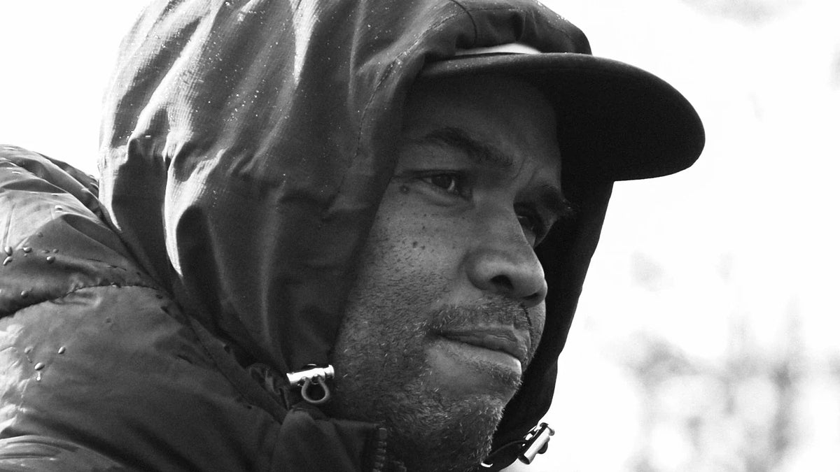 A Sharks coach in a black hooded puffer jacket and cap, beaded with rain, watches a training session with autumn trees behind him, in black and white.