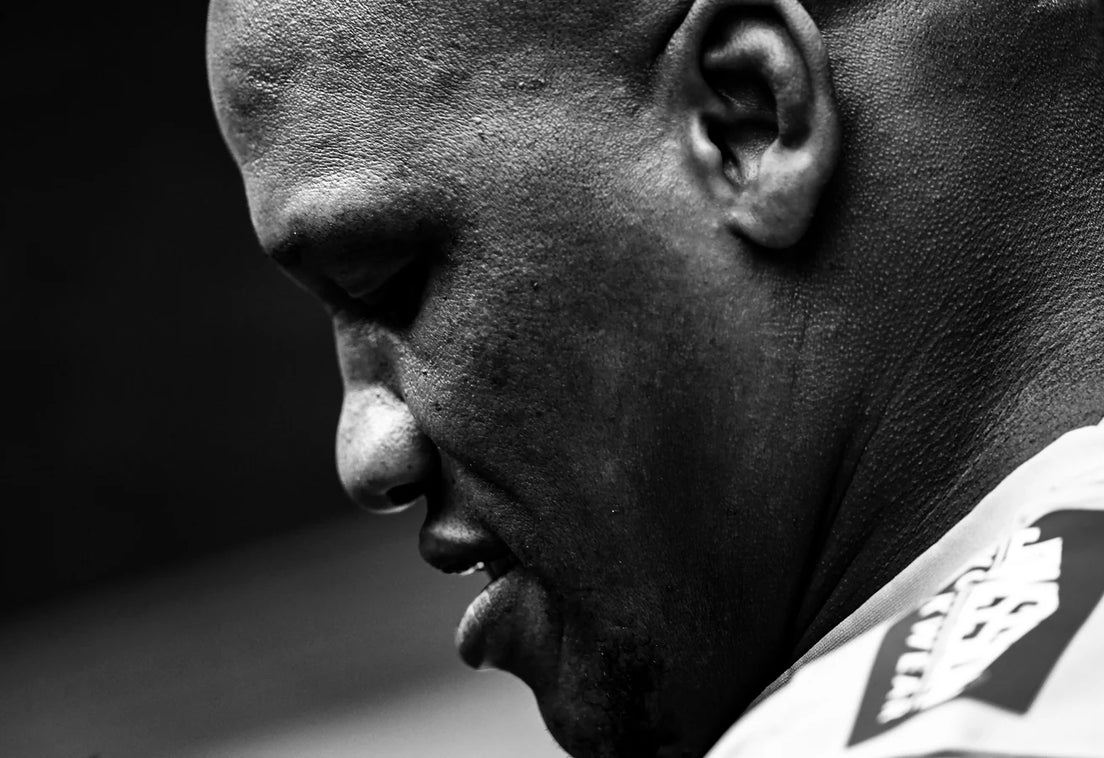 Black and white close-up of Bongi Mbonambi in profile, head bowed and eyes lowered, wearing a white Sharks training top.