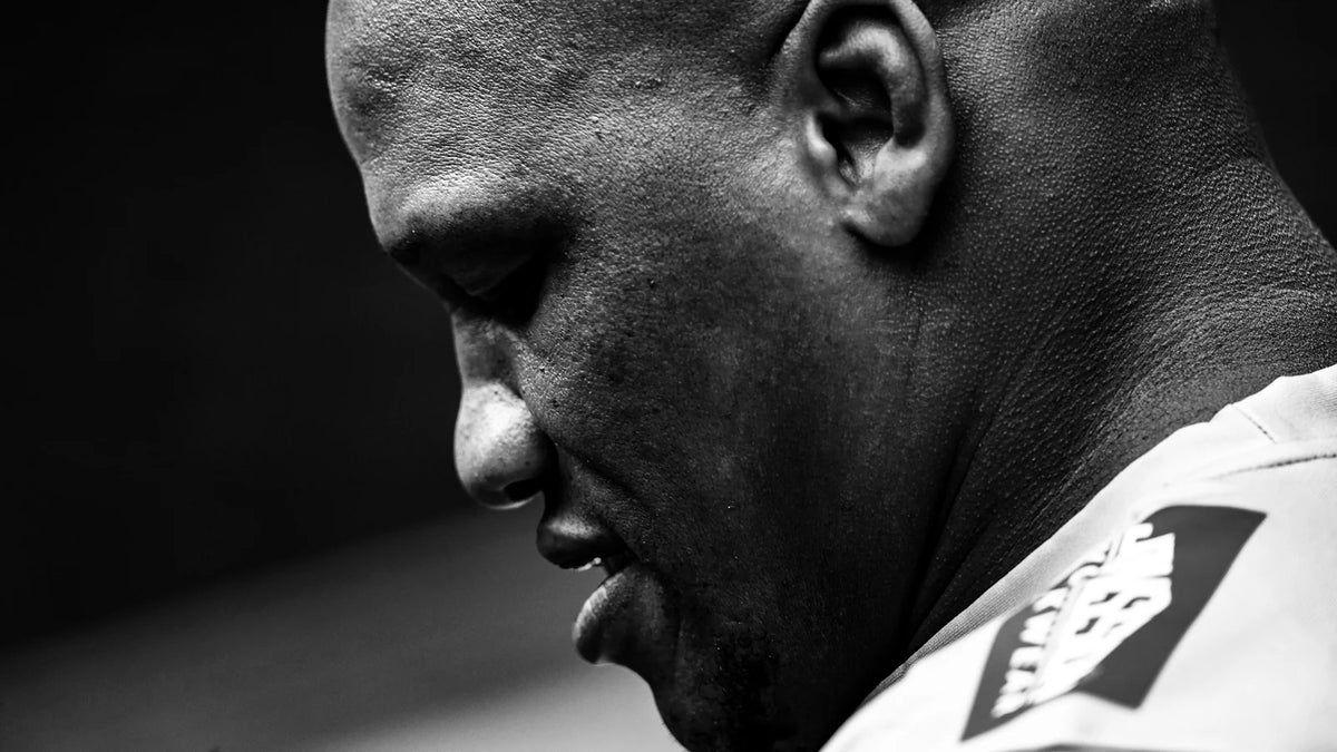 Black and white close-up of Bongi Mbonambi in profile, head bowed and eyes lowered, wearing a white Sharks training top.