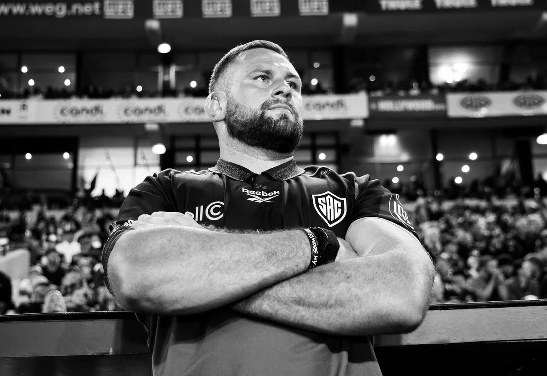 A Hollywoodbets Sharks player in a black warm-up top stands arms folded at the touchline, looking up, with a packed stand blurred behind him.