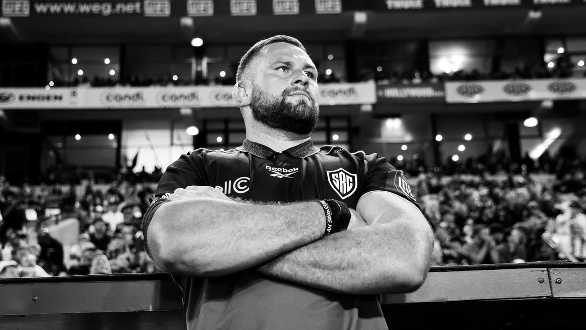 A Hollywoodbets Sharks player in a black warm-up top stands arms folded at the touchline, looking up, with a packed stand blurred behind him.