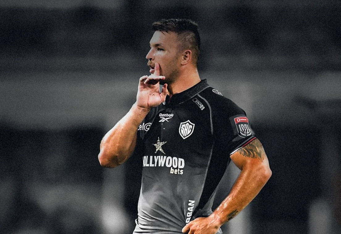 A Hollywoodbets Sharks player holds a finger to his lips with his other hand on his hip, picked out in colour against a blurred black and white stadium background.