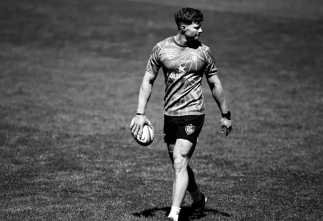 A Sharks player in a training shirt walks across a sunlit field holding a rugby ball at his side and glancing over his shoulder, in black and white.