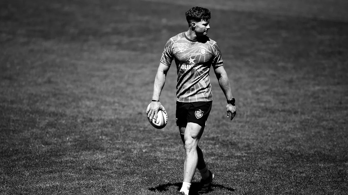 A Sharks player in a training shirt walks across a sunlit field holding a rugby ball at his side and glancing over his shoulder, in black and white.