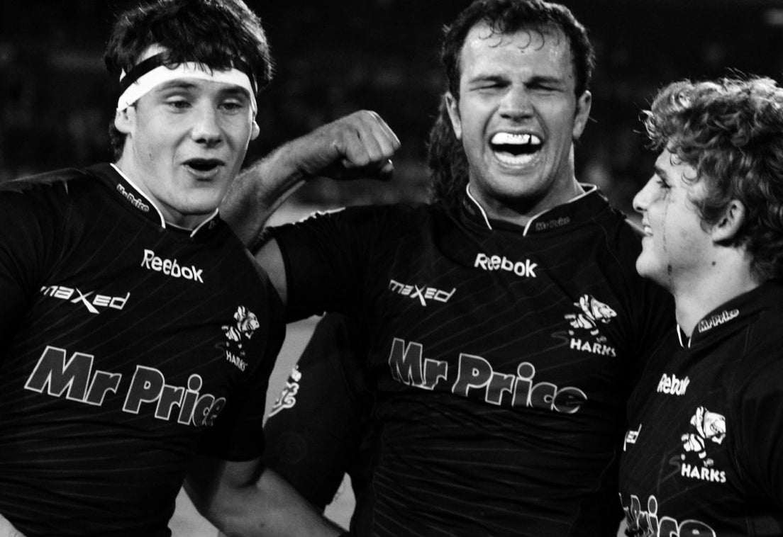 Three Sharks players in black Super Rugby-era jerseys celebrate together on the pitch at night, the player in the centre laughing with his fist raised.