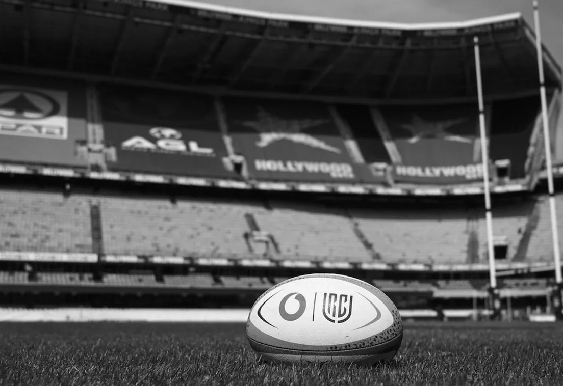 Black and white photo of a Vodacom URC match ball on the grass at an empty Hollywoodbets Kings Park, with the stands and goalposts behind.