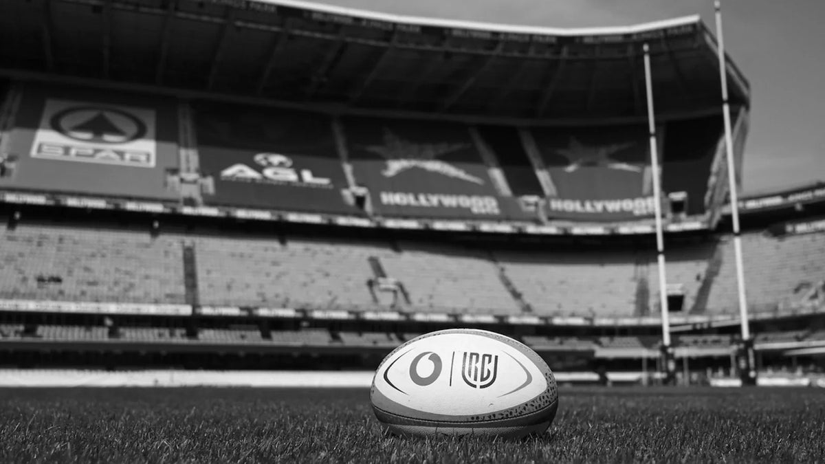 Black and white photo of a Vodacom URC match ball on the grass at an empty Hollywoodbets Kings Park, with the stands and goalposts behind.