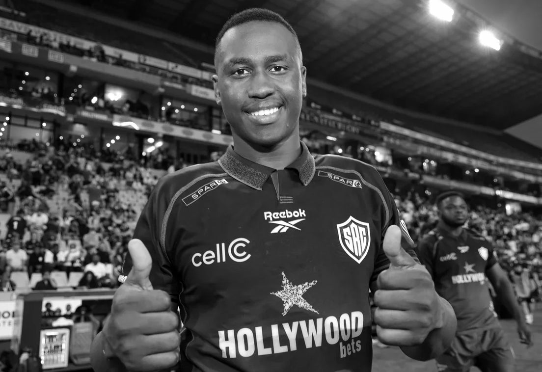 Vusi Moyo smiles and gives two thumbs up in a Hollywoodbets Sharks jersey on the pitch, with floodlit stands and spectators behind him, in black and white.