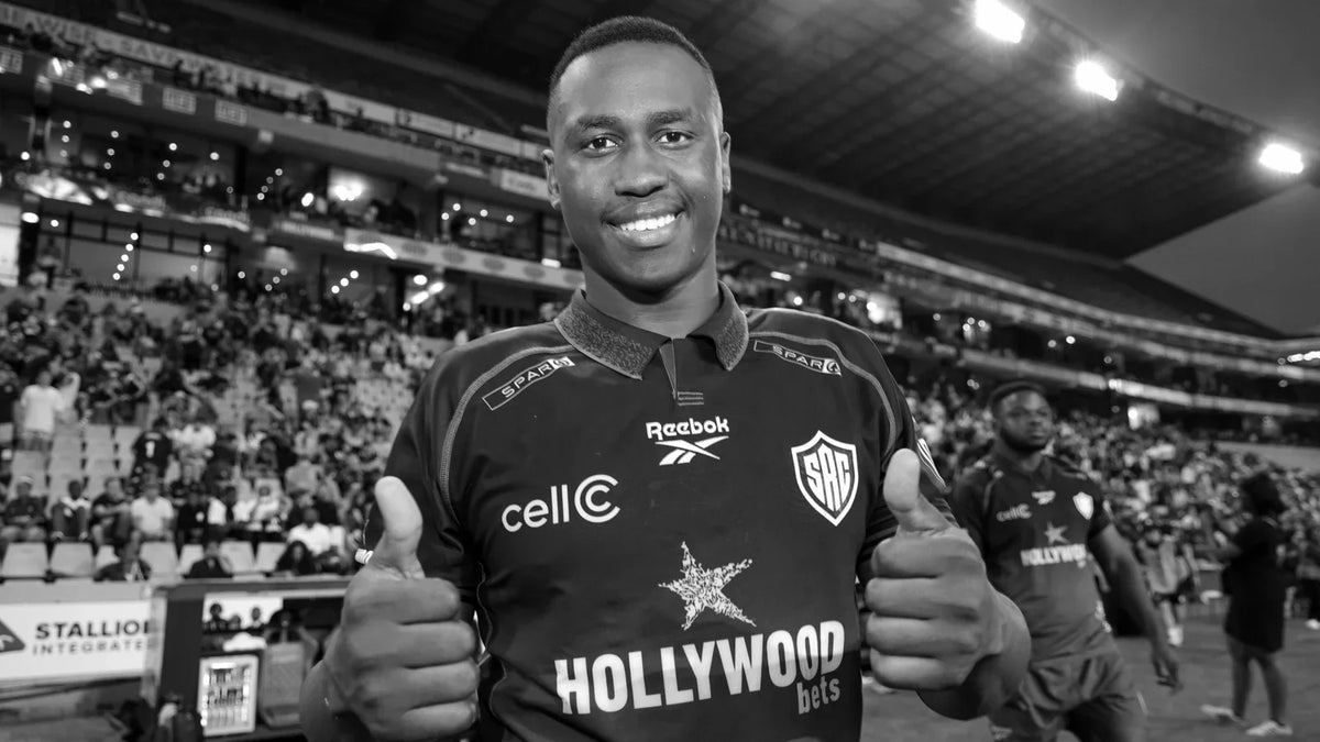 Vusi Moyo smiles and gives two thumbs up in a Hollywoodbets Sharks jersey on the pitch, with floodlit stands and spectators behind him, in black and white.