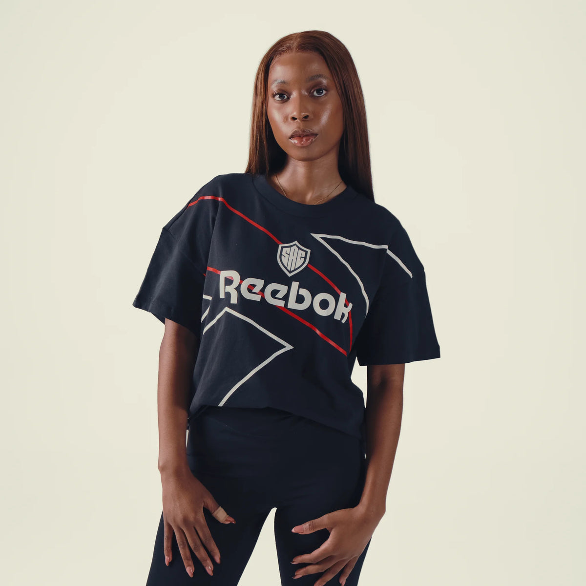A woman modeling a dark navy blue Reebok short-sleeve t-shirt with white and red graphic details and the Reebok logo on the front, set against a solid light beige background.