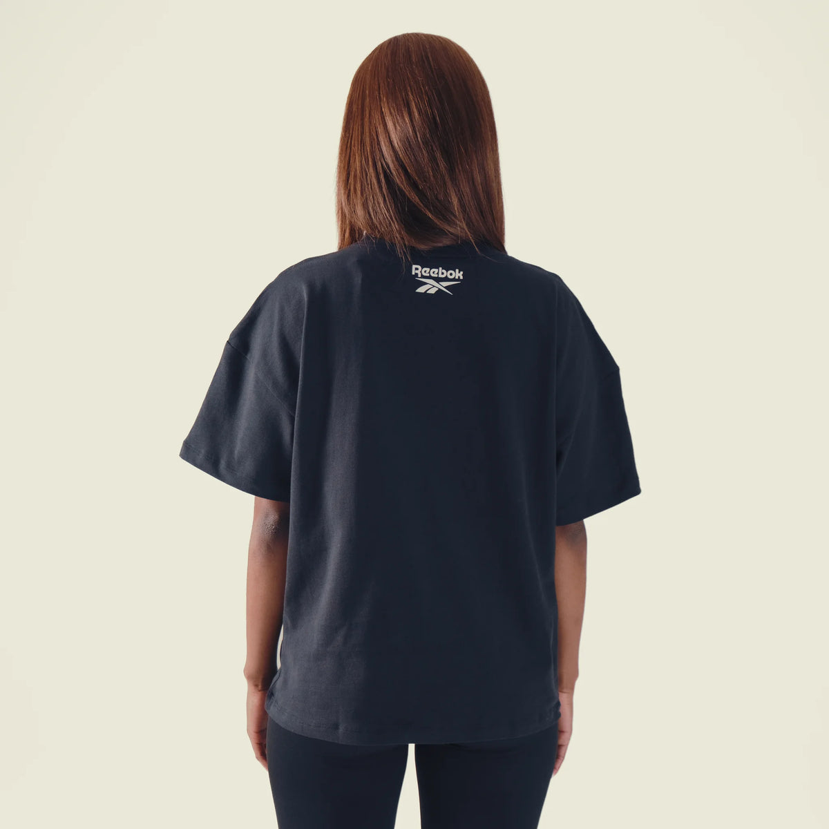 A woman viewed from behind wearing a dark navy blue short-sleeve t-shirt with a white Reebok logo printed on the upper back, set against a solid light beige background.