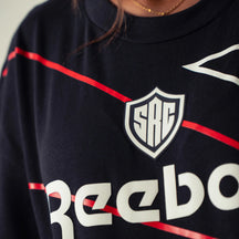 Close-up of a person wearing a dark navy tshirt with a white and red Reebok shield logo, a white Reebok wordmark across the chest.