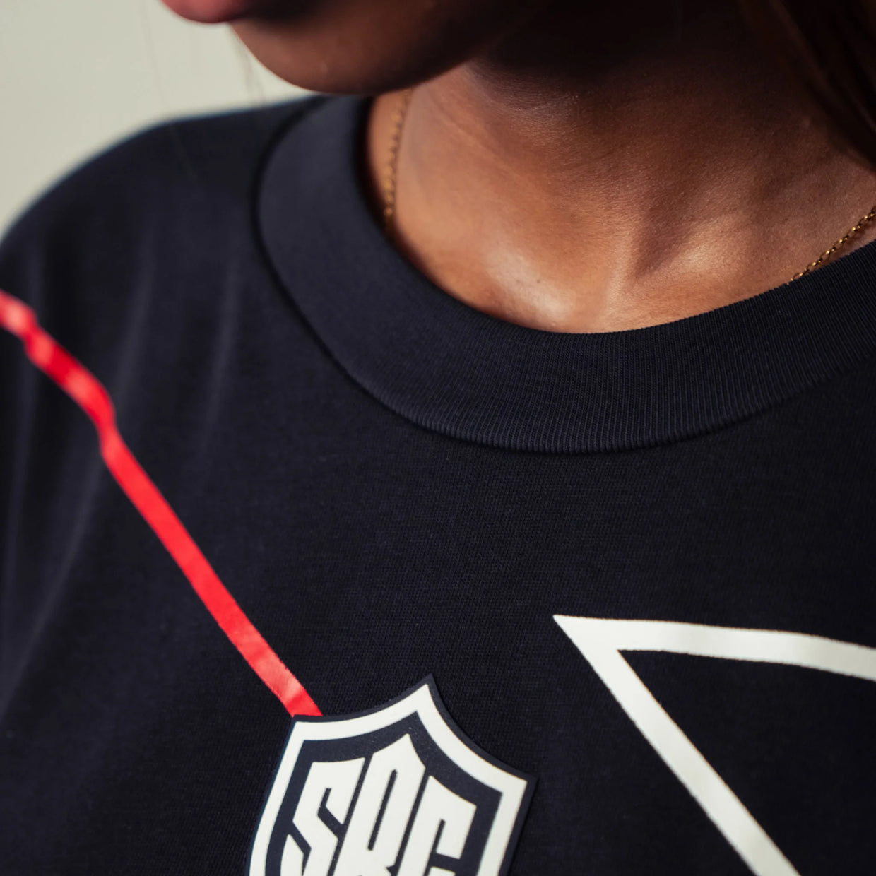 Close-up of a person wearing a dark navy crewneck sweatshirt with a white shield logo patch and a red line detail on the chest.