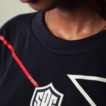 Close-up of a person wearing a dark navy crewneck sweatshirt with a white shield logo patch and a red line detail on the chest.