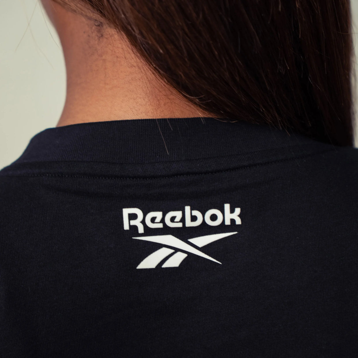 Close-up of the back of a person wearing a black Reebok t-shirt, showing the white Reebok logo and vector crossmark printed on the upper back.